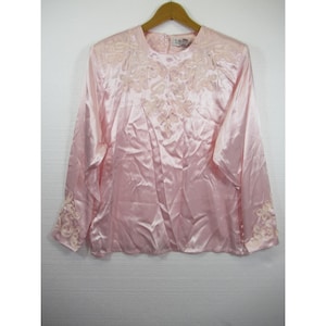 May include: A light pink satin blouse with long sleeves, featuring delicate embroidered floral details on the shoulders and cuffs. The blouse is hanging on a wooden hanger against a white background.