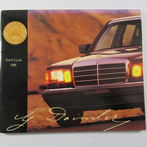May include: A brochure for the 1986 Mercedes-Benz S-Class. The cover features a partial view of the car's front, with the headlights illuminated. The Mercedes-Benz logo is visible, along with the text "The S-Class 1986" and a signature.