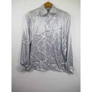 May include: A silver-colored button-down shirt with long sleeves and a collared neckline. The shirt is made of a shiny, possibly satin-like material, and is hanging on a wooden hanger.