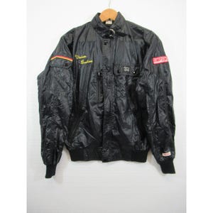 May include: Black vintage racing jacket with a stand-up collar, featuring the text "Vivian Burton" in gold script on the chest and a "Simpson" patch. The jacket has multiple pockets, a ribbed hem, and cuffs. The jacket is hanging on a wooden hanger.