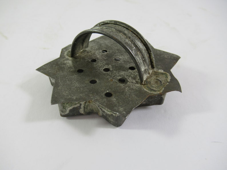 May include: A vintage metal cookie cutter shaped like a star with a handle. The cutter has a worn, gray finish and multiple holes throughout.