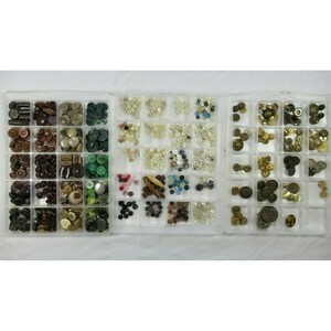 May include: A collection of various buttons in a clear plastic organizer. The buttons are in different colors, shapes, and sizes, including brown, green, white, and gold. Some buttons have multiple holes, while others have shanks.