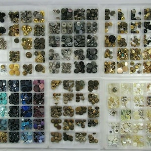 May include: A collection of assorted buttons in various colors and materials, organized in a clear plastic storage box. The buttons are arranged in rows and columns, with different types of buttons in each compartment. The buttons include plastic, metal, and fabric buttons in a variety of shapes, sizes, and colors.