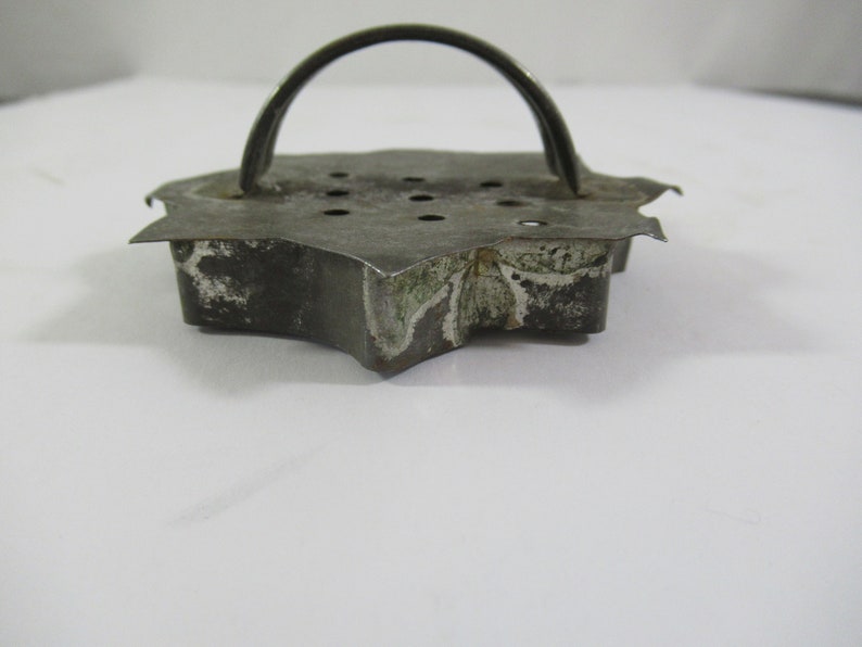 May include: A vintage metal cookie cutter shaped like a star with a handle. The cutter has a worn, rustic look with some rust spots and a faded design.