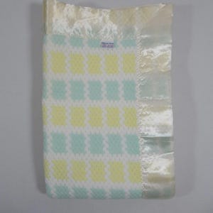 May include: A baby blanket with a repeating pattern of yellow and light blue squares on a white background. The blanket has a satin border in a cream color. A small label is visible in the upper left corner.