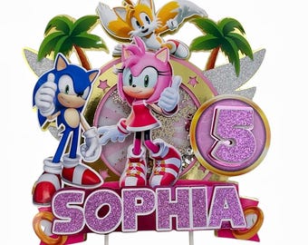 Op de afbeelding: Een roze, gouden en blauwe Sonic the Hedgehog-taarttopper met de naam "Sophia" en het nummer "5" erop. De topper toont Sonic, Tails en Amy Rose.