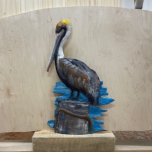 May include: A carved wooden pelican sculpture with a brown body, white head, and long beak. The bird is perched on a wooden piling with blue accents, and a rope detail. The sculpture is set against a light wood background.