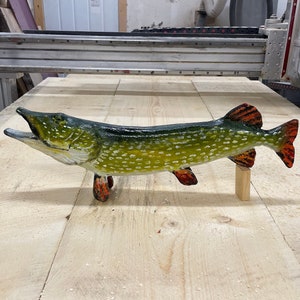 May include: A hand-carved wooden fish sculpture of a pike, painted in realistic detail with green, yellow, and orange colors. The fish is mounted on a wooden stand.