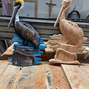 May include: Two wooden pelican sculptures. One is painted with brown, black, and yellow details, perched on a blue base. The other is unpainted, showing the natural wood grain. Both have rope accents.