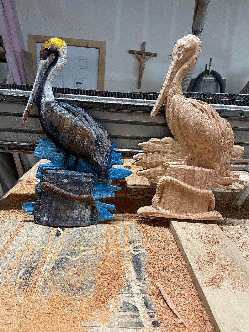 May include: Two wooden pelican sculptures. One is painted with brown, black, and yellow details, perched on a blue base. The other is unpainted, showing the natural wood grain. Both have rope accents.