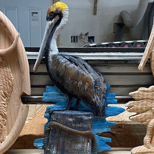 May include: A hand-carved wooden pelican sculpture, painted in shades of brown, gray, and blue. The bird is perched on a wooden piling with blue water accents. The sculpture is a decorative item.
