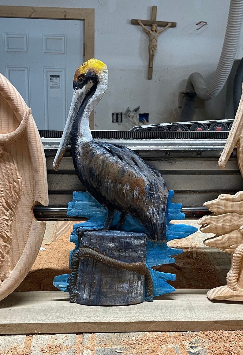 May include: A hand-carved wooden pelican sculpture, painted in shades of brown, gray, and blue. The bird is perched on a wooden piling with blue water accents. The sculpture is a decorative item.