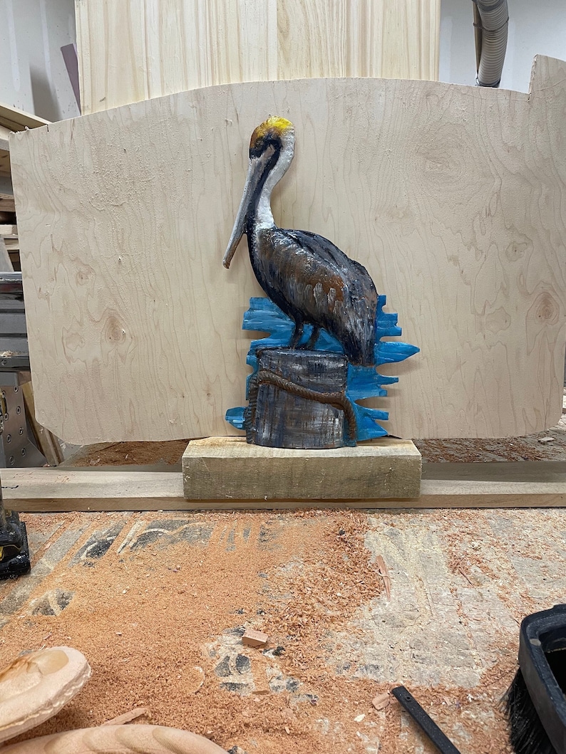 May include: A carved wooden pelican sculpture with a brown body, white head, and long beak. The bird is perched on a wooden piling with blue accents, and a rope detail. The sculpture is set against a light wood background.