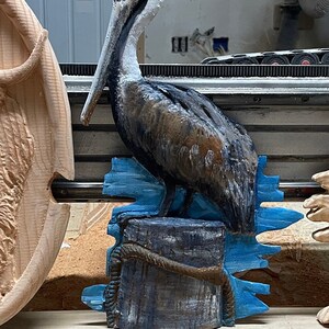 May include: A carved wooden pelican sculpture. The bird has a yellow head, a long white beak, and brown and black feathers. It is perched on a blue and gray wooden post with rope detailing, with blue water-like accents.