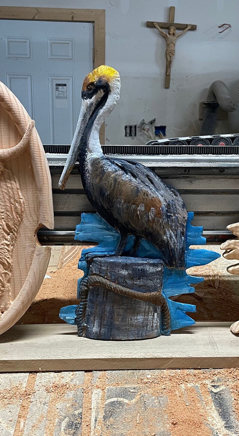May include: A carved wooden pelican sculpture. The bird has a yellow head, a long white beak, and brown and black feathers. It is perched on a blue and gray wooden post with rope detailing, with blue water-like accents.