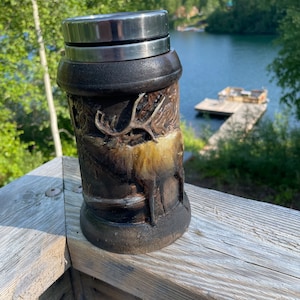Puede incluir: Una taza de viaje marrón y negra con una tapa de acero inoxidable. La taza tiene un diseño de un ciervo en un entorno de bosque.