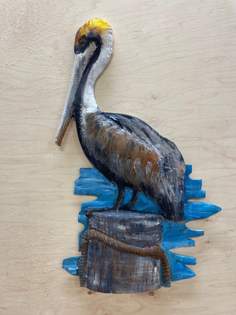 May include: A hand-painted wooden pelican sculpture. The bird is perched on a weathered wooden piling, with blue water and rope details. The pelican has brown, gray, and white feathers, with a yellow-gold head and beak.