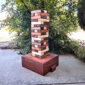 May include: A large wooden tower game with a base. The tower is made of blocks in various shades of brown, red, and natural wood. The base is a dark brown wooden box with a small step on the side.