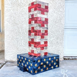 May include: A large, red, white, and blue wooden tower game with a base that has a blue and gold star pattern. The tower game is made of wooden blocks that are stacked on top of each other.
