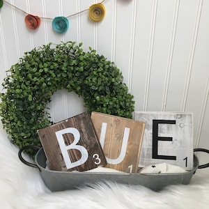 Puede incluir: Tres fichas de Scrabble de madera que deletrean la palabra "BUE" en letras blancas. Las fichas est&aacute;n en una bandeja de metal con una corona de laurel verde y una guirnalda de flores de papel en el fondo.