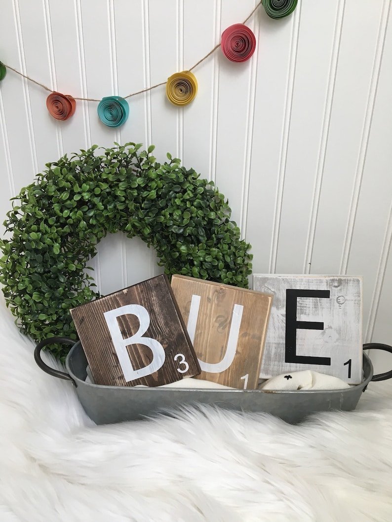Puede incluir: Tres fichas de Scrabble de madera que deletrean la palabra "BUE" en letras blancas. Las fichas est&aacute;n en una bandeja de metal con una corona de laurel verde y una guirnalda de flores de papel en el fondo.
