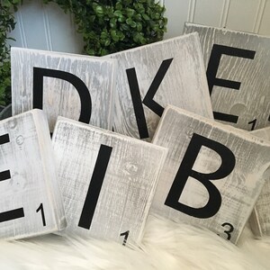 Puede incluir: Azulejos de madera blancos con letras negras que deletrean la palabra "DEIBAK" con los valores de las letras en la parte inferior de cada azulejo.