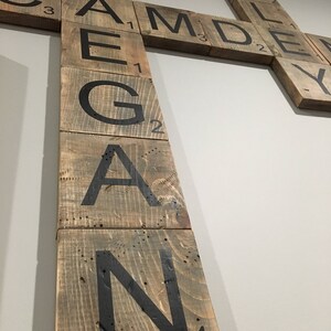 Puede incluir: Una pieza de arte de pared de madera hecha de fichas de Scrabble que deletrean la palabra "REAGAN". Las fichas est&aacute;n dispuestas verticalmente y est&aacute;n pintadas de marr&oacute;n con letras negras. Las fichas tienen los valores de puntos para cada letra impresos en ellas.