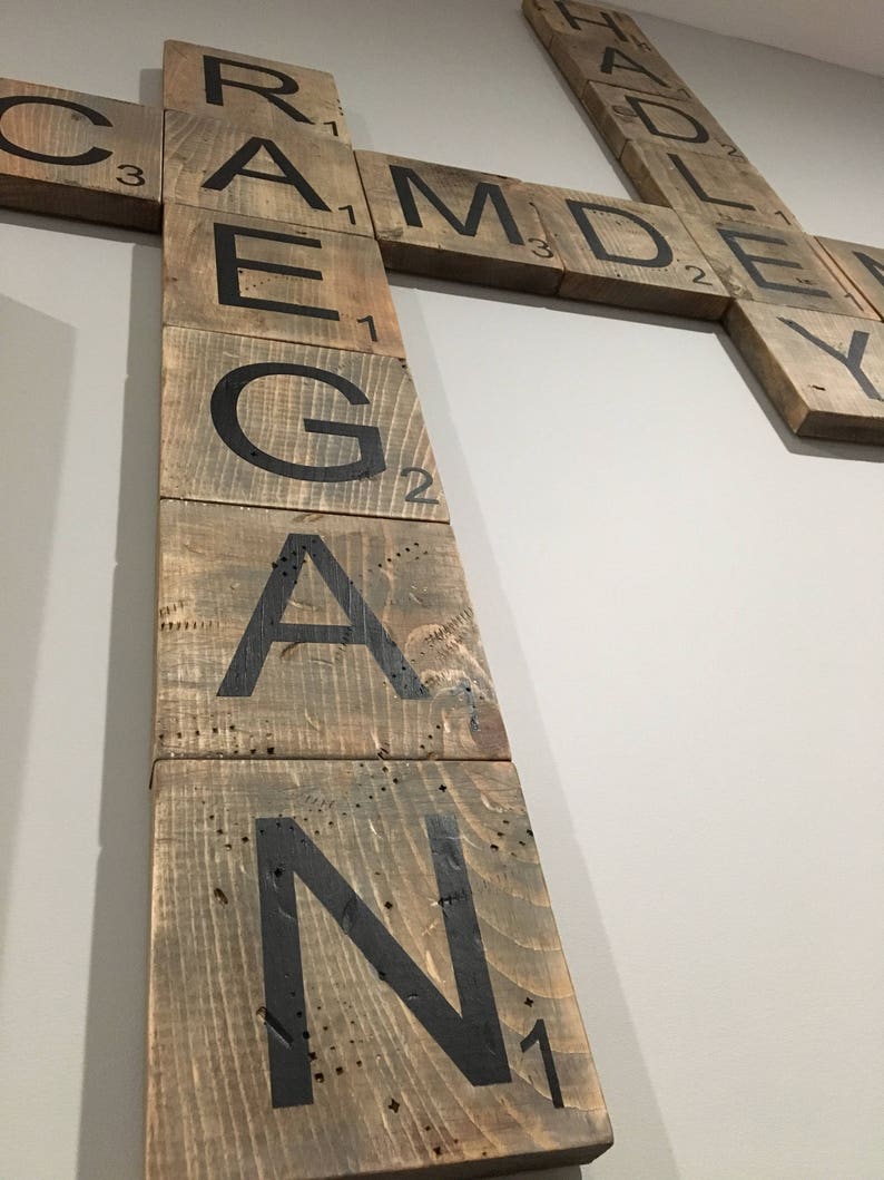 Puede incluir: Una pieza de arte de pared de madera hecha de fichas de Scrabble que deletrean la palabra "REAGAN". Las fichas est&aacute;n dispuestas verticalmente y est&aacute;n pintadas de marr&oacute;n con letras negras. Las fichas tienen los valores de puntos para cada letra impresos en ellas.