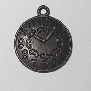 May include: A round, bronze-colored clock charm with a loop at the top. The clock face features stylized numbers and ornate detailing. The hands point to approximately 1:07. This charm is suitable for jewelry making or crafting.