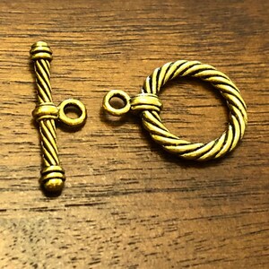 May include: A gold-toned metal toggle clasp with a twisted rope design. The clasp has a bar with a loop on one end and a ring on the other end.