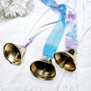 May include: Three brass bells with a gold finish are attached to sheer ribbons. The ribbons are blue and white, with butterfly patterns in purple and blue. The bells are on a white textured surface.