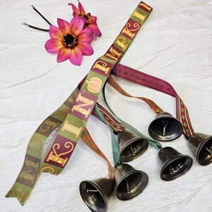 May include: A decorative ribbon featuring the text PUMPKIN in yellow letters on a green and maroon background, accented with a small orange pumpkin icon. Five dark metallic bells are attached to the ends of the ribbon, accompanied by two pink flowers.