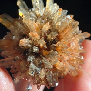 May include: Close-up of a cluster of golden-brown crystals with sharp, pointed edges. The crystals have a translucent quality, with hints of orange and brown throughout. The cluster is held in a person's hand against a dark background.