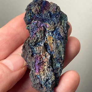 Puede incluir: Un espécimen mineral oscuro y texturizado con colores iridiscentes de púrpura, azul y oro. La superficie rugosa de la roca refleja la luz, creando un efecto brillante. El mineral se sostiene en una mano.