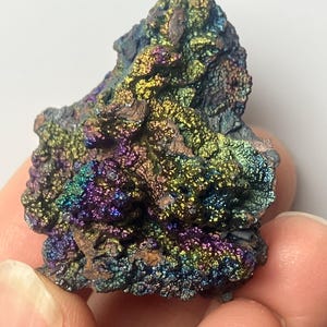 May include: A colorful, iridescent mineral specimen. The rock displays a variety of colors, including purple, green, gold, and blue. The surface has a textured, rough appearance. The mineral is held between two fingers.