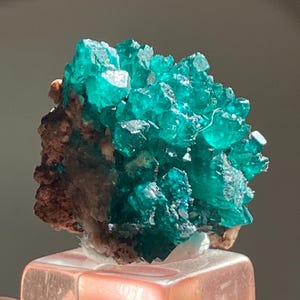 May include: A vibrant, crystalline mineral specimen featuring a cluster of bright teal-green crystals. The crystals are clustered on a matrix of brown and beige rock. The specimen is mounted on a clear, rectangular base.