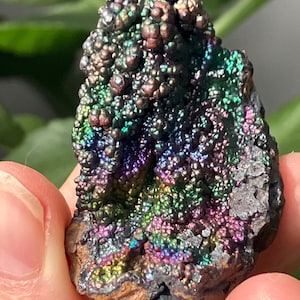 Puede incluir: Primer plano de un espécimen mineral con una superficie vibrante e iridiscente. El mineral muestra una gama de colores, incluyendo verdes, azules, púrpuras y dorados. La textura parece burbujeante y cristalina, con una base oscura y rugosa.