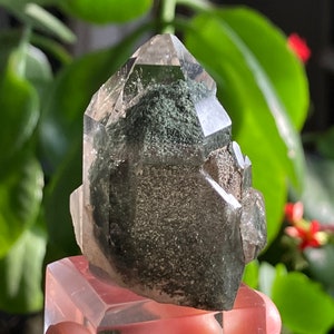 May include: A clear quartz crystal with green inclusions. The crystal is faceted and has a pointed tip. It is sitting on a clear acrylic base.