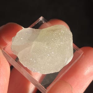 May include: A light-colored, crystalline mineral specimen with a sparkly texture, resting on a clear, square acrylic base. The mineral has a rough, irregular shape and is held by fingers against a dark background.