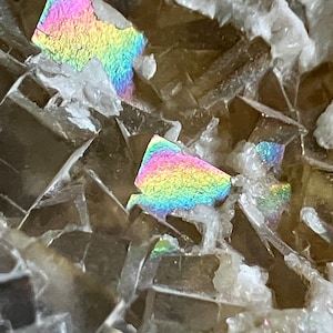 May include: Close-up of a brown and gray iridescent mineral with a rainbow sheen. The mineral has a crystalline structure and is composed of many small, flat crystals.