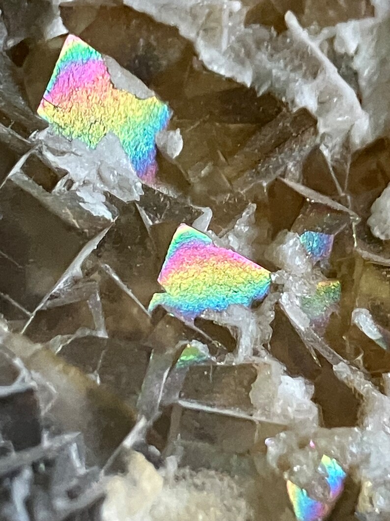 May include: Close-up of a brown and gray iridescent mineral with a rainbow sheen. The mineral has a crystalline structure and is composed of many small, flat crystals.