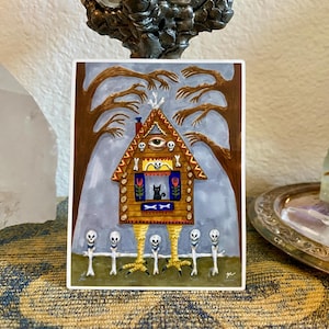 May include: A whimsical illustration of a house with chicken legs, a third eye, and a black cat in the window. The artwork features a spooky theme with a fence of skull-topped posts and bare trees in the background. The color palette includes brown, yellow, and gray.