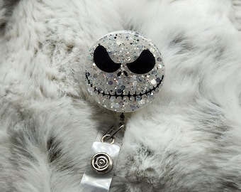May include: A white glitter badge reel featuring a black and white Jack Skellington face design. The reel is attached to a clear plastic badge holder.
