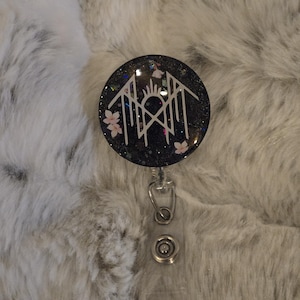 May include: A black glitter badge reel with a white geometric design, pink flowers, and a sunburst. The reel has a clear plastic strap and a metal clip. The background is a fluffy, white and gray surface.