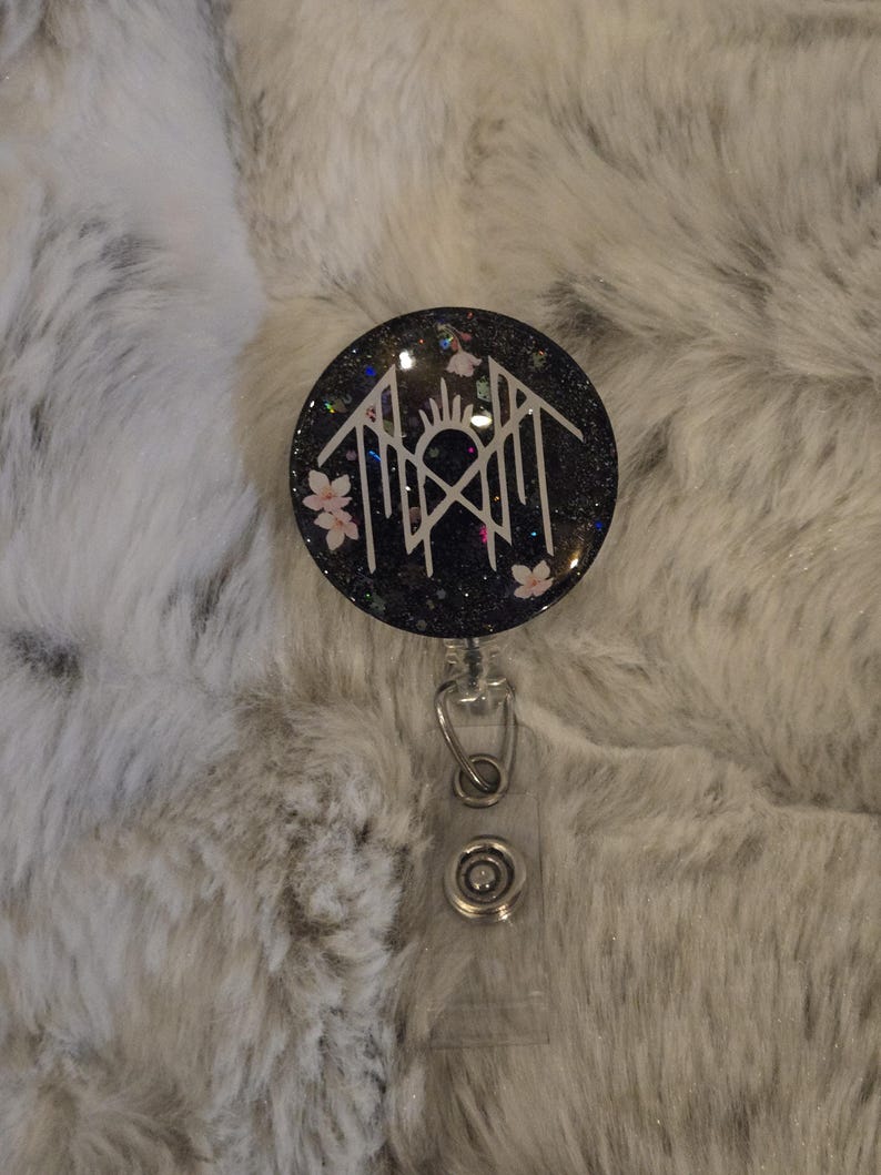 May include: A black glitter badge reel with a white geometric design, pink flowers, and a sunburst. The reel has a clear plastic strap and a metal clip. The background is a fluffy, white and gray surface.