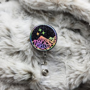 May include: A black glitter badge reel with a holographic mountain and star design. The reel is attached to a clear plastic retractable badge holder.