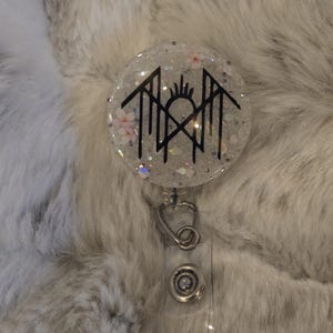 Peut inclure: Un enrouleur de badge rond et transparent avec un fond blanc scintillant. L'enrouleur de badge présente un motif graphique noir avec des accents floraux. L'enrouleur a un clip en métal argenté et une sangle en plastique transparent.
