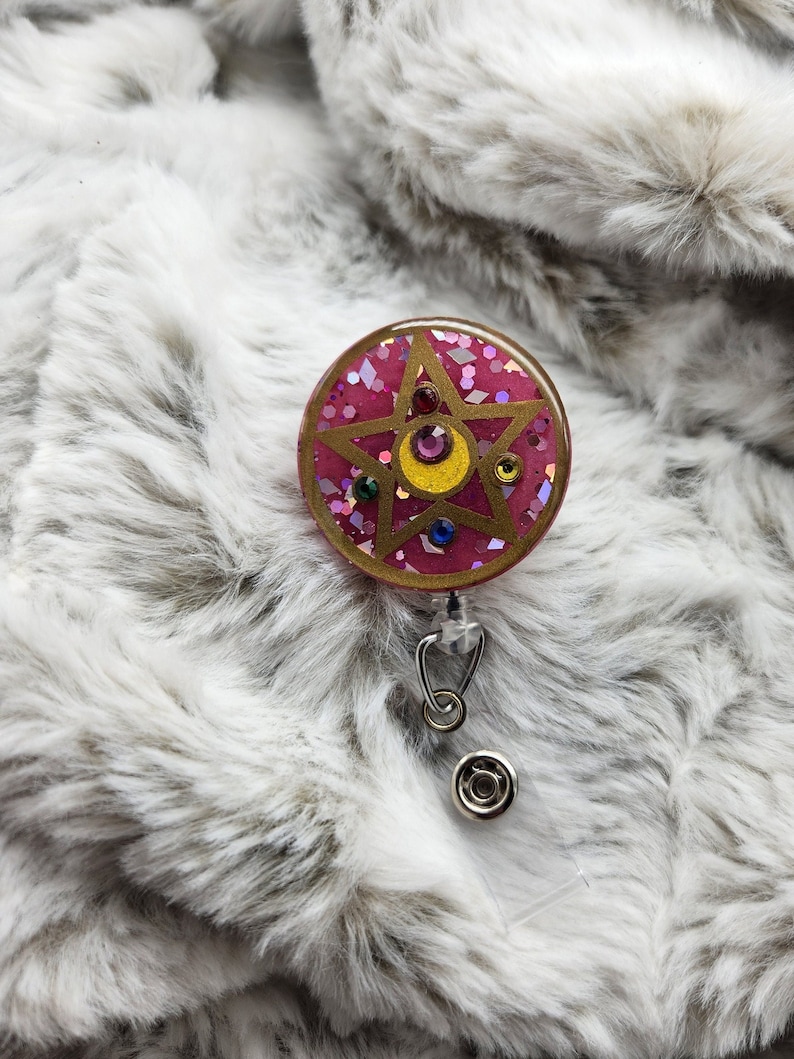 May include: A pink and gold glitter resin badge holder with a five-pointed star and a crescent moon design. The star has a yellow center and is surrounded by smaller, colorful glitter.