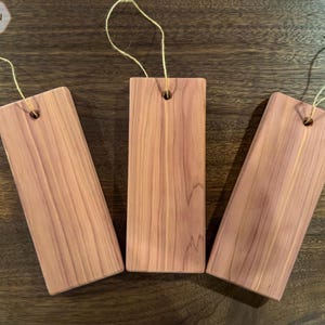 May include: Three rectangular cedar wood blocks with a hole at the top for hanging. Each block has a natural wood grain pattern and is tied with a thin, light-colored string. The blocks are arranged on a dark wood surface.