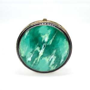 May include: A round, silver-toned compact with a green and white marbled design. The compact has a hinged lid and a black rim.